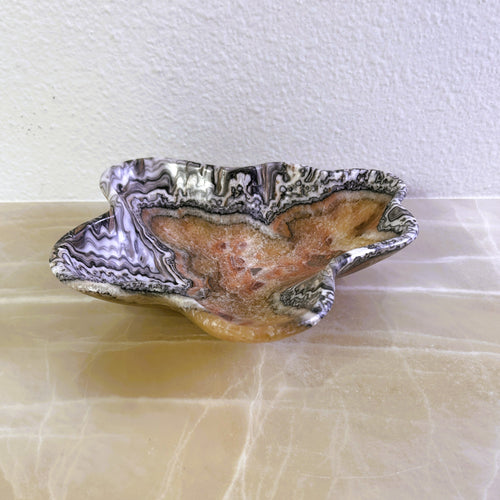 An elegant hand-carved stone bowl with capricious veins in beige and light brown tones, placed on a marble surface.